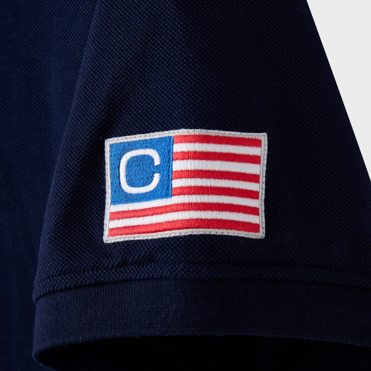 HMDD Glo Navy Polo, close-up of the sleeve patch, a woven stars-and-stripes flag with a white C in the blue canton