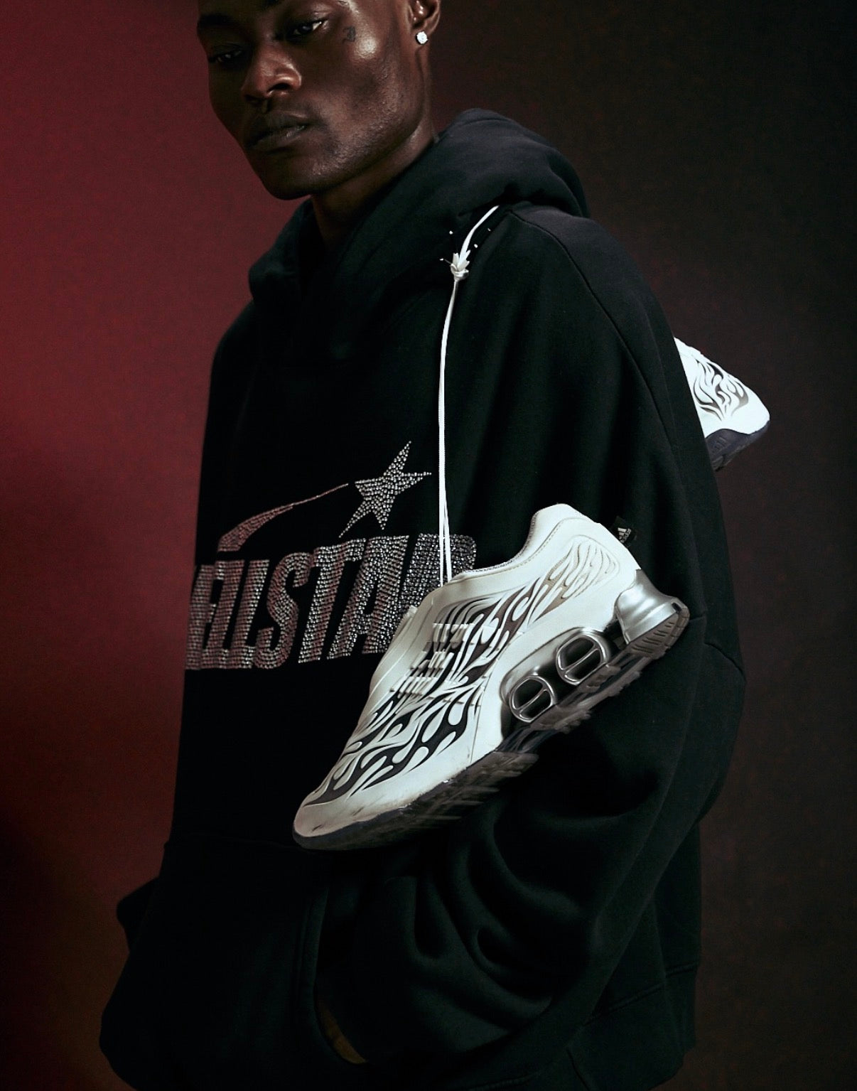 Hellstar Adidas Megaride White, model in a black Hellstar hoodie holding the pair by the laces against a dark red backdrop