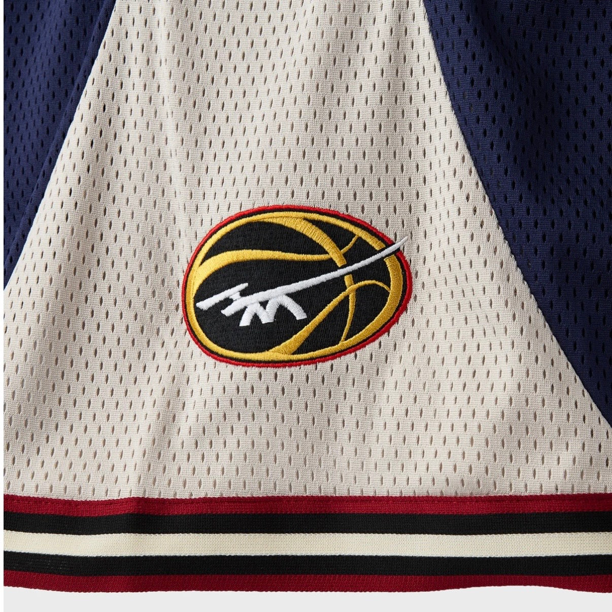 HMDD UE Basketball Shorts, close-up of the gold and maroon basketball logo on cream mesh above the striped hem