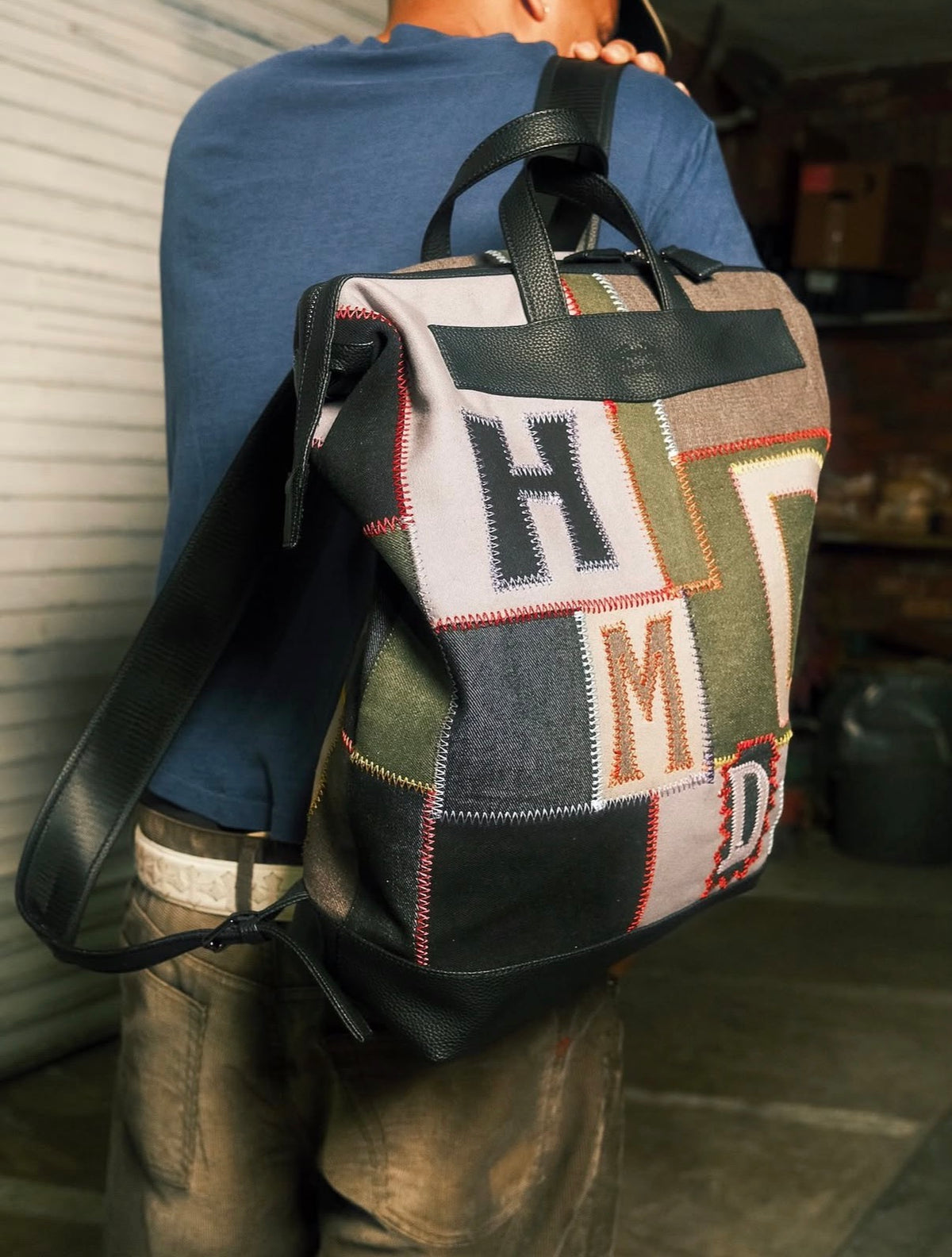 Model wearing the HMDD Hopscotch Backpack on one shoulder, showing the patchwork H, M and D panels and black leather trim