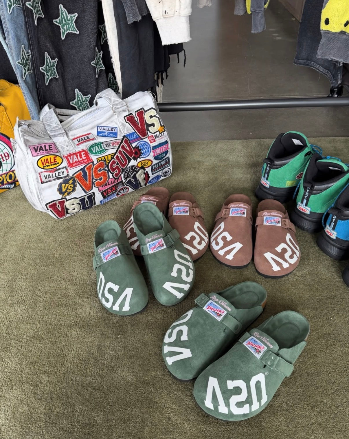 Vale Airport Duffle 1/100, in-store shot of the duffle on green carpet beside rows of green and brown suede clogs
