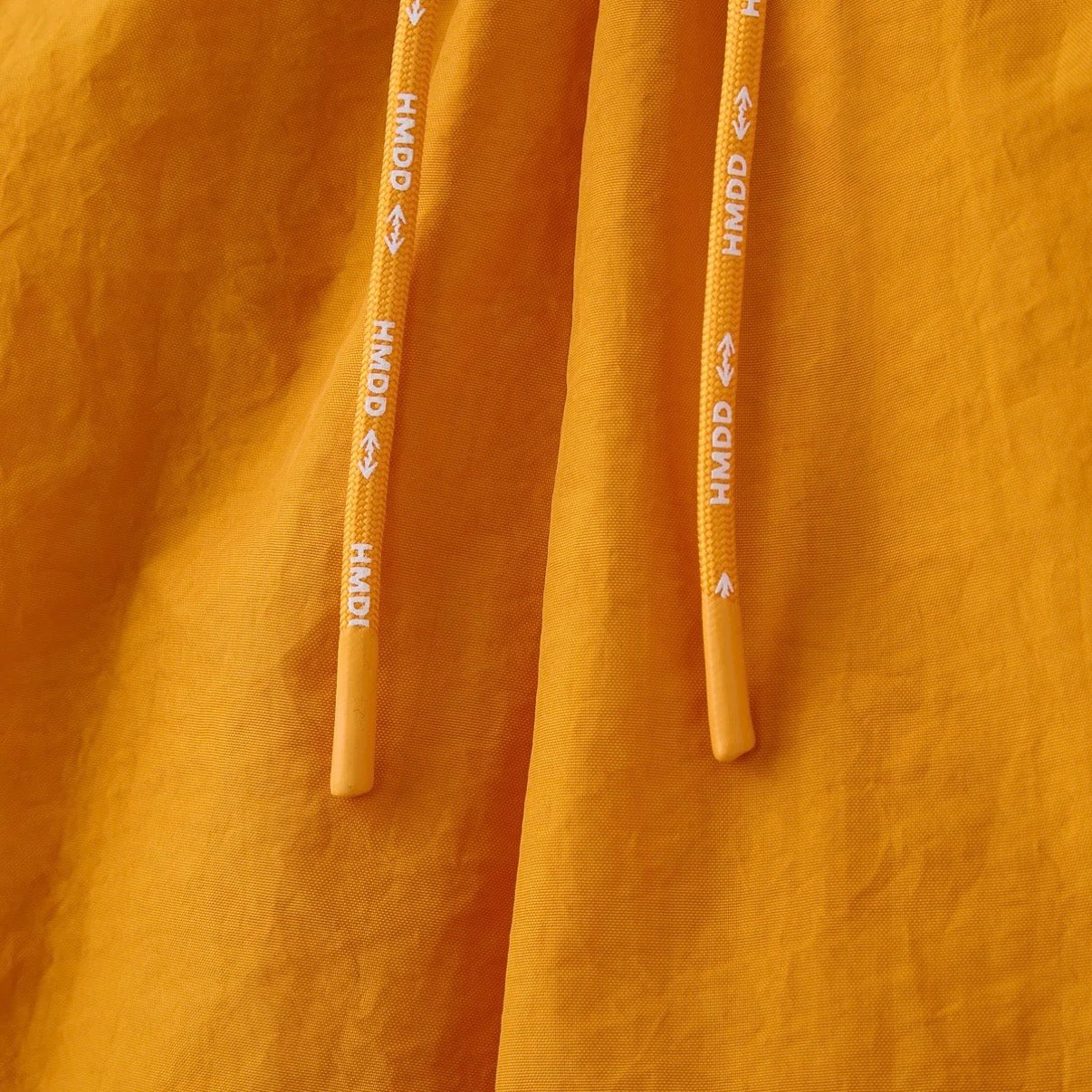 HMDD Gold Juggernaut 3/4 Shorts, close-up of the two drawcord ends printed with HMDD against the orange fabric