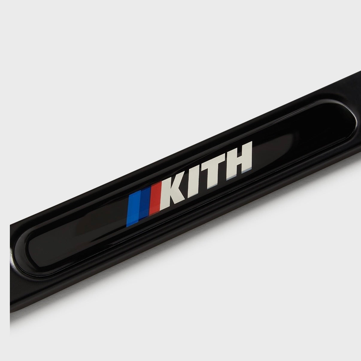 Kith BMW Black License Plate Holder, close-up of the lower bar, white Kith wordmark and blue and red stripes on gloss black