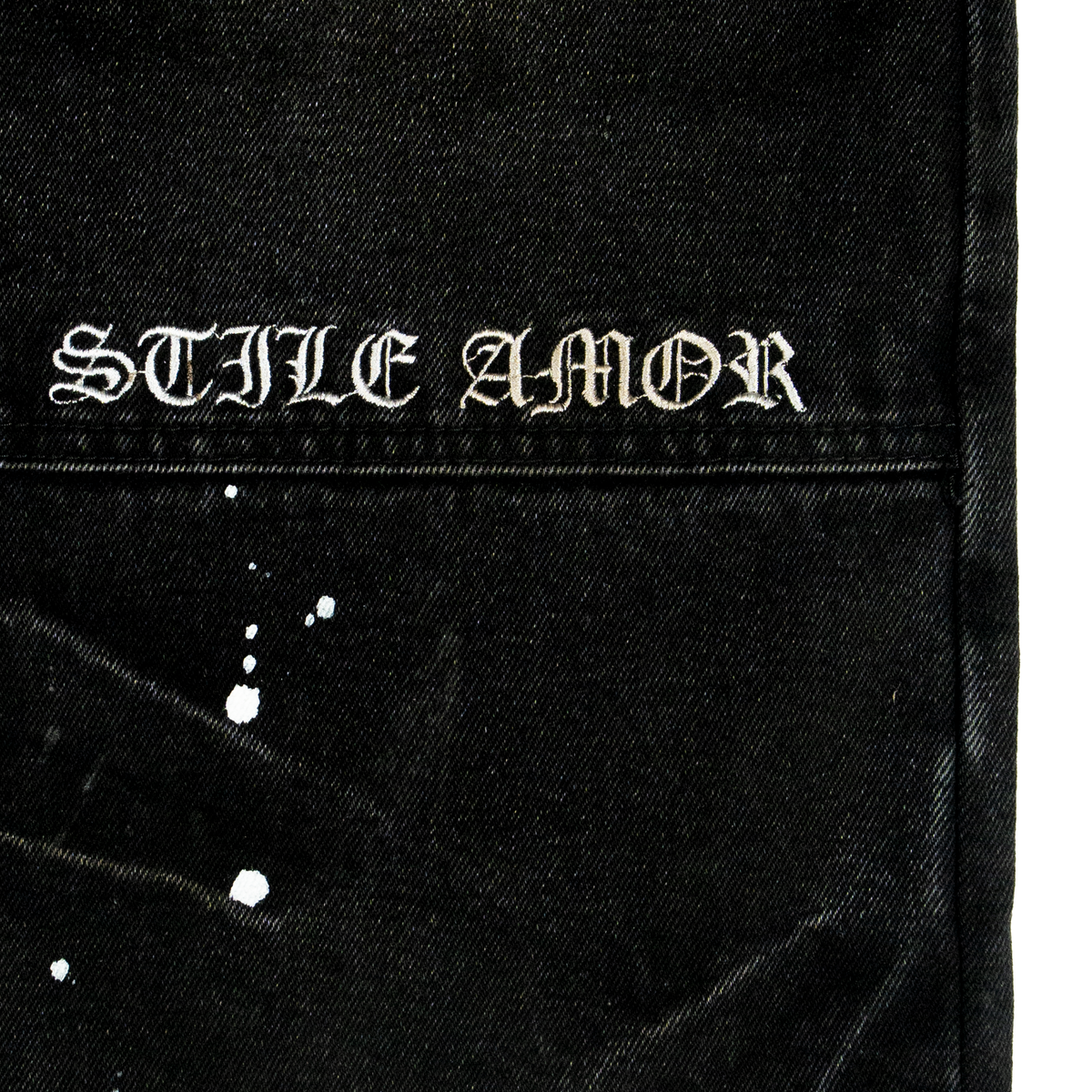 Close-up of white embroidered STILE AMOR gothic lettering and paint flecks on black denim