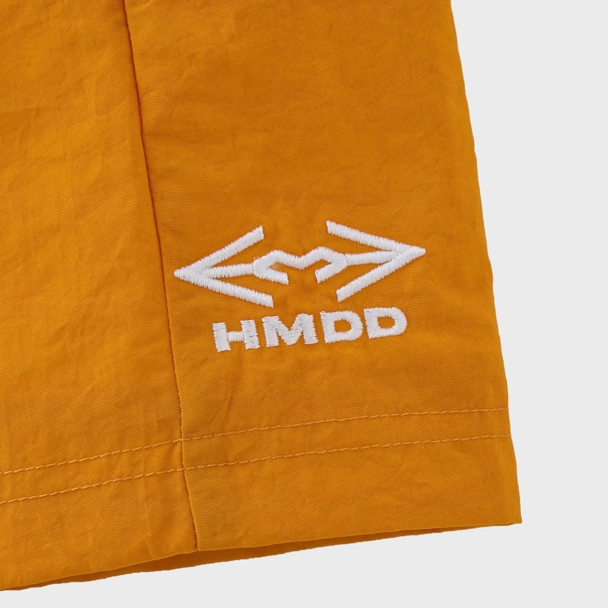 HMDD Gold Juggernaut 3/4 Shorts, close-up of the large white winged HMDD logo printed on the orange leg panel