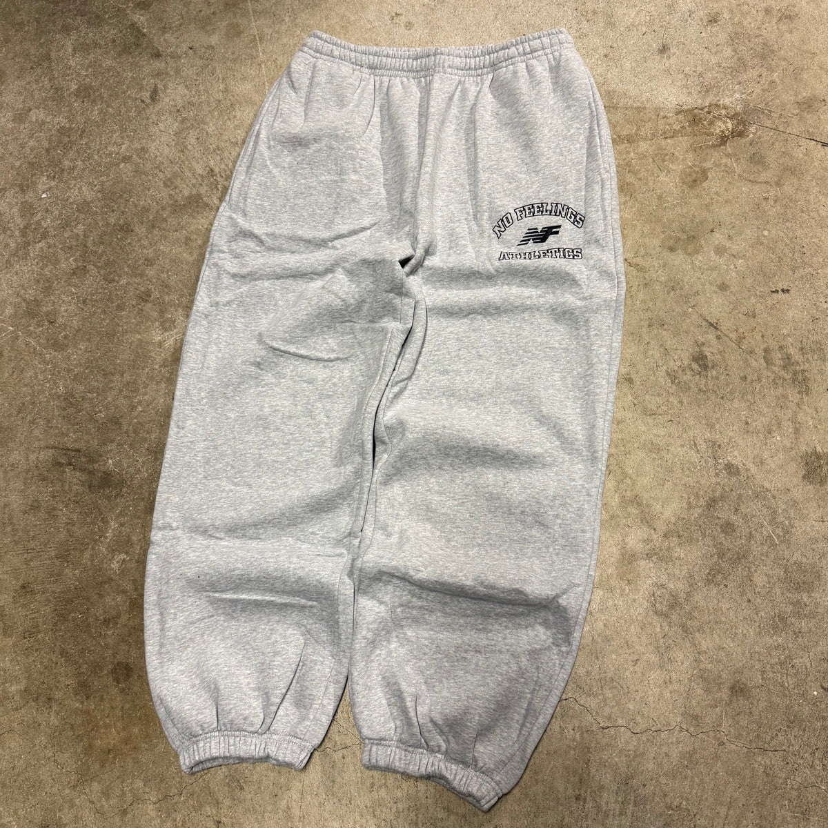 No Feelings heather grey sweatpants laid flat, with a navy athletics graphic on the upper thigh and elastic cuffs