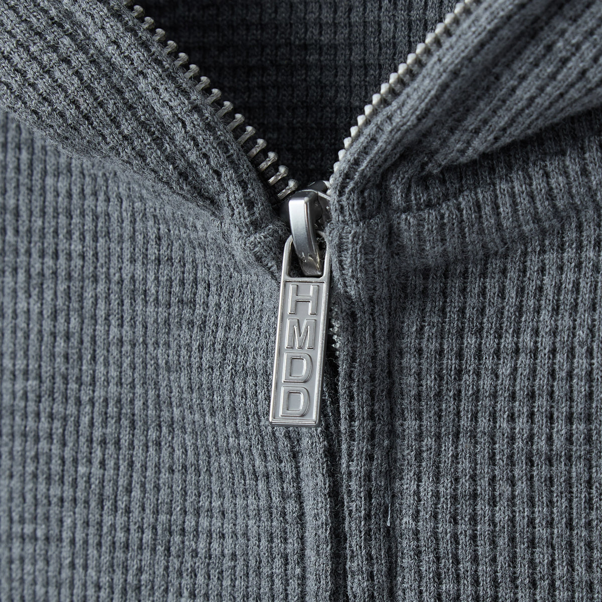 Close-up of the hoodie zip pull engraved HMDD on grey waffle thermal fabric